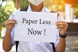 Thai boy student holds protesting paper which has text, Paper Less Now, soft and selective focus, concept for calling teachers and students to reduce paper using at school to save our environment. photo