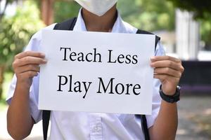 Thai boy student holds protesting paper which has text 'Teach Less Play More', soft and selective focus, concept for calling all teachers to reduce teaching in class and increase playing outclass . photo