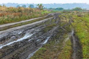 Mañana neblinosa otoñal paisaje rústico con camino de tierra en primer plano y pequeños edificios en la colina al fondo foto