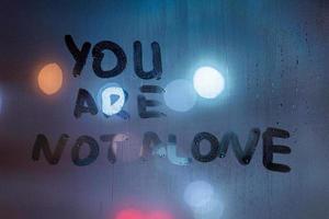 the phrase you are not alone written by finger on night wet glass with blurred classic blue lights in background photo