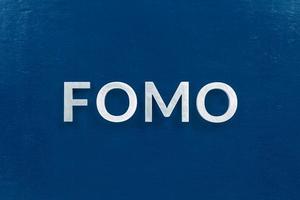 la palabra abreviada fomo - miedo a perderse - colocada con letras plateadas en una superficie plana de color azul clásico foto