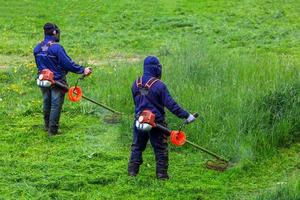 two lawnmower men with string trimmer and face mask trimmong grass - close-up photo