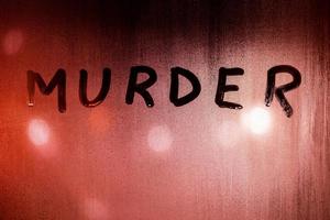 the word murder handwritten on wet glass of night window with bloody red back light of street lights photo