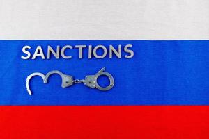 the word sanctions laid with silver metal letters on russian tricolor flag near handcuffs in directly above view photo