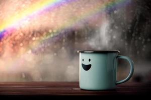 felicidad y mente positiva, concepto de salud mental. disfrutando del café con dibujos animados de cara sonriente, lluvia borrosa con arco iris como vista exterior. sonríe en un día lluvioso foto