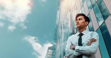 Portrait of a Striving Young Asian Businessman in the City. Crossed Arms and looking up into the Sky. Modern Office Building as background photo