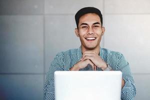 feliz joven hombre de negocios asiático trabajando en una computadora portátil en su lugar de trabajo. manos en la barbilla, sonriendo y mirando a la cámara foto