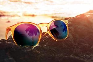 cierre de gafas de sol con reflejo de palmera de coco en la isla, puesta de sol como fondo, tono cálido y vintage, viajar por la playa en verano o proteger los ojos del concepto de luz solar foto