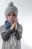 niña molesta con un sombrero gris envuelto en una bufanda sobre un fondo blanco, esperando el frío invierno. foto