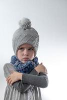 Upset little girl in a gray hat wrapped in a scarf on a white background, waiting for the cold winter. photo