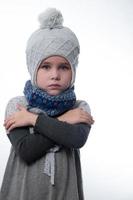 Upset little girl in a gray hat wrapped in a scarf on a white background, waiting for the cold winter. photo