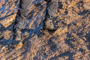 Piernas en pantalones grises y botas de montaña caminando hacia arriba en una colina fangosa a la luz del sol de la tarde foto