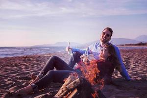 pareja joven sentada en la playa al lado de una fogata bebiendo cerveza foto