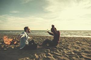 pareja joven sentada en la playa al lado de una fogata bebiendo cerveza foto