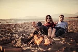 pareja joven sentada en la playa al lado de una fogata bebiendo cerveza foto