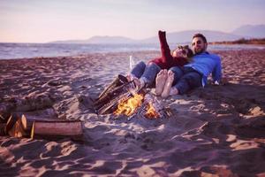 pareja joven sentada en la playa al lado de una fogata bebiendo cerveza foto