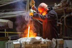 worker with hard hat and face mask stirs liquid metal in a furnace by steel bar photo