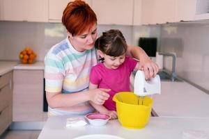 Funny little girl helper playing with dough on his hands learning to knead helps adult mom in the kitchen, happy cute baby daughter and parent mom have fun cooking cookies. photo