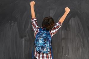 boy celebrates a successful schooling with his hands in the air as he faces the school blackboard photo