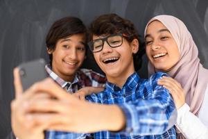 Group of arab teens taking selfie photo on smart phone with black chalkboard in background. Selective focus