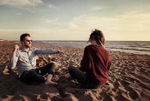 pareja joven sentada en la playa al lado de una fogata bebiendo cerveza foto