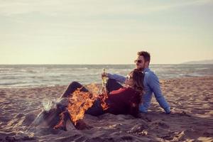 pareja joven sentada en la playa al lado de una fogata bebiendo cerveza foto