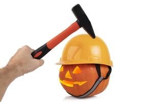 Orange festive pumpkin with glowing eyes in the form of a lantern, halloween, in a yellow construction helmet and a man's hand with a hammer. Security concept on holidays. photo