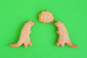 galletas de mantequilla caseras en forma de dinosaurios con una nube de texto con texto - ¿cómo estás? - sobre fondo verde, vista superior. foto