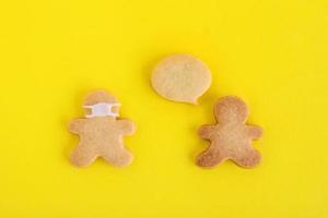 Homemade cookies in shapes of people with callout cloud and with face medical mask on yellow background, top view. Sweet shortbread with white glaze. Social distancing concept. photo