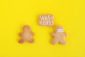 Galletas caseras de pan dulce con glaseado blanco sobre fondo amarillo, vista superior. dos hombres, uno de ellos con máscara facial y con una nube de llamadas con texto - lavarse las manos. foto