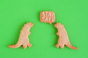 Two homemade cookies in shapes of dinosaurs with inscription - Stay safe - on green background, top view. Sweet shortbread with white glaze. Social distancing concept. photo