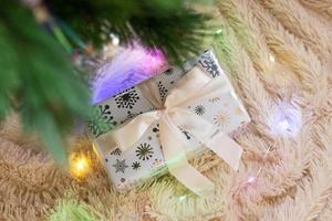 Top view on Christmas gift wrapped on white paper with ribbon on a background of beige plush plaid with Christmas lights, under Christmas tree. photo