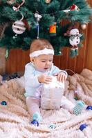 Cute little girl is sitting near Christmas tree on a beige plush plaid with Christmas decorations and lights and try to open Christmas gift. photo