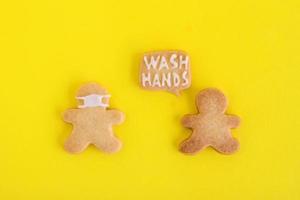 Homemade cookies in shapes of people with inscription - Wash hands - and with face medical mask on yellow background, top view. Sweet shortbread with white glaze. Social distancing concept. photo