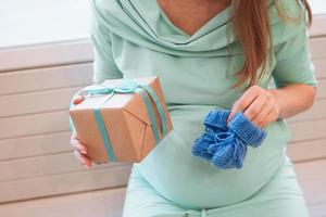 Pregnant woman in blue dress is holding gift box and blue knitted bootee. Pregnant lifestyle. photo