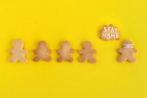 Homemade cookies in shapes of people with inscription - Stay home - and with face medical mask on yellow background, top view. Sweet shortbread with white glaze. Social distancing concept. photo