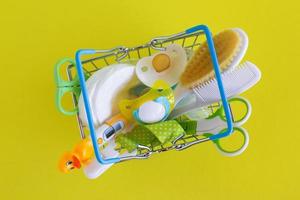 Flat lay on shopping basket with baby care items - scissors, hairbrushes, pacifiers, thermometer, cotton pads, pacifier holders and nasal aspirator - on yellow background. photo