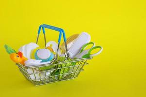 Shopping basket with baby care items - scissors, hairbrushes, pacifiers, thermometer, cotton pads, pacifier holders and nasal aspirator - on yellow background. photo