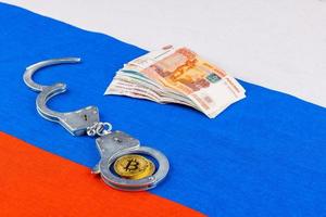 bunch of russian paper rubles currency and bitcoin shiner chained with handcuffs on russian flag background photo