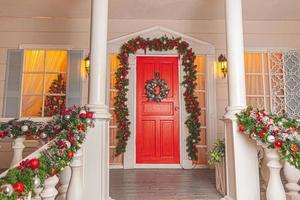Christmas porch decoration idea. House entrance with red door decorated for holidays. Red and green wreath garland of fir tree branches and lights on railing. Christmas eve at home photo