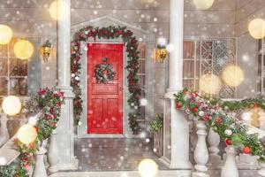 Christmas porch decoration idea. House entrance with red door decorated for holidays. Red and green wreath garland of fir tree branches and lights on railing. Christmas eve at home photo