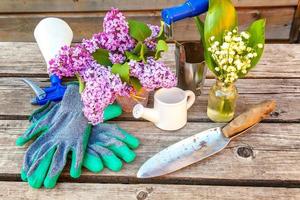 Gardening tools, watering can, shovel, spade, pruner, rake, glove, lilac, lily of the valley flowers on vintage wooden table. Spring or summer in garden, eco, nature, horticulture hobby concept. photo