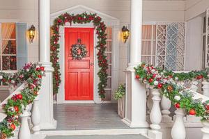 Christmas porch decoration idea. House entrance with red door decorated for holidays. Red and green wreath garland of fir tree branches and lights on railing. Christmas eve at home photo