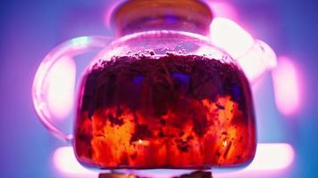 bule transparente com folhas de chá preto ferve em um queimador de gás contra uma luz de fundo roxa neon. bolhas borradas em água quente fervendo dentro de uma chaleira de vidro. video