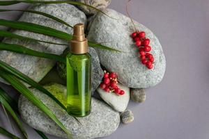 A bottle of green color with a spray bottle lies on the stones, next to the green leaves and berries of schisandra chinensis photo