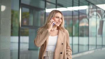 glückliche blonde frau in einem braunen mantel geht und spricht am telefon. Schöne Geschäftsfrau kommuniziert auf einem Smartphone, während sie an einem Herbsttag durch die Stadt geht. Zeitlupe.