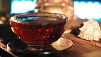 Black Hot Tea Poured into Beautiful Glass against background of the Smoke Coming from Scented Sticks, backlit by Warm Light. Tea ceremony. Oriental Cup close up. Unrecognizable person. Slow motion video