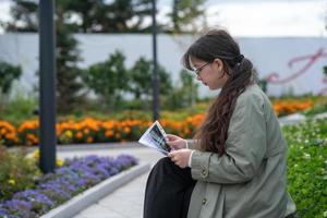 A girl sits on a bench on the street with documents in her hands and using her phone photo