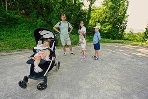 Family with three kids walking at park, baby girl hold toy in sitting stroller. photo