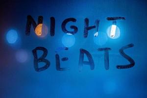 the words night beats handwritten on wet window glass at nighttime with blue light color photo